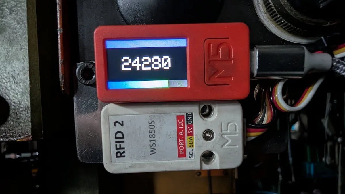 機械のフレームに白いテープで留められたM5StickC PlusとRFID 2ユニット。画面には打刻数の24280が表示されている